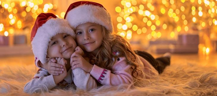 Děti ležící na koberci ve svitu vánočního osvětlení. Na hlavě mají červené vánoční čepice. 